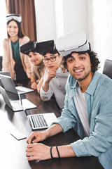 Wall Mural - smiling group of multiethnic business partners with virtual reality headsets at table with laptops in modern office