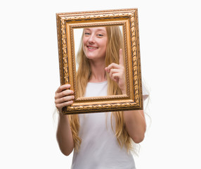 Sticker - Blonde teenager woman holding vintage frame art surprised with an idea or question pointing finger with happy face, number one