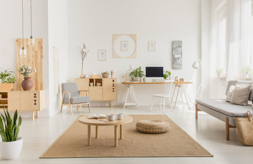 Real photo of a modern home office interior with wooden furniture, coffee table, sofa, pouf and computer on a desk