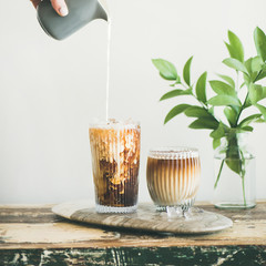 Poster - Iced coffee in tall glasses with milk poured over from pitcher by hand, white wall and green plant branches at background, copy space, square crop. Summer refreshing beverage ice coffee concept