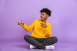 © Drobot Dean - Portrait of stylish african american guy smiling and pointing fingers aside at copyspace while sitting on floor with legs crossed, isolated over violet background