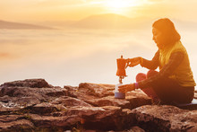 A Woman Traveler Boils A Geyser Coffee Machine And Drinks A Hot Drink From The Mug, Admiring The Colorful Dawn With The Fog In The Mountains, Camping And Hiking In Nature Concept
