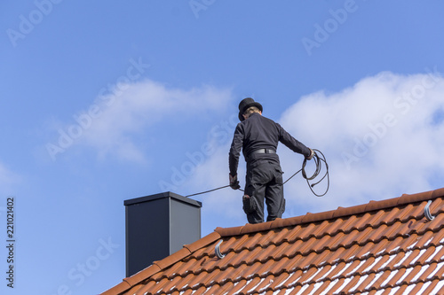 Fotomural Chimney sweep cleaning a chimney standing on the house roof, lowering equipment