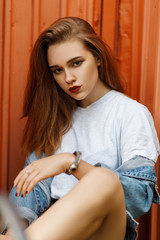 Canvas Print - Stylish young model woman in a gray T-shirt and denim vintage jacket sits near an orange metal wall