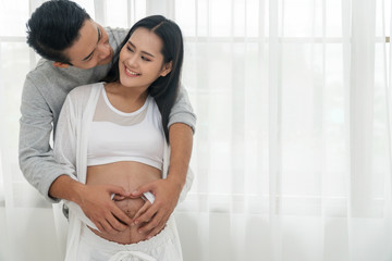 Husband embracing his wife making heart shape on the pregnant belly with their hands. Happy couple of young man hugs his pregnant beautiful wife, smiling and looking into each other's eyes at home.