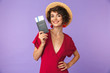 © Drobot Dean - Photo of charming young woman 20s wearing big straw hat smiling while holding passport and travel tickets, isolated over white background