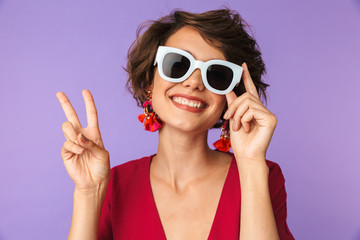 Canvas Print - Image of fashionable brunette woman 20s in straw hat and sunglasses showing peace sign, isolated over violet background