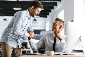 Wall Mural - image of happy businessmen coworkers 30s in formal clothes working in office, and shaking hands toge
