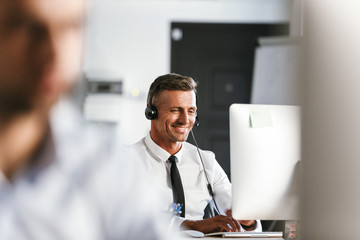 Wall Mural - photo of businesslike man 30s wearing office clothes and headset, smiling while sitting by computer 