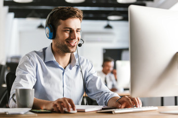 Wall Mural - Photo of young worker man 20s wearing office clothes and headset, smiling and talking with clients in call center