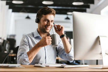 Wall Mural - photo of businesslike man 20s wearing office clothes and headset, drinking tea from cup while sittin