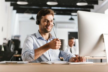 Wall Mural - photo of businessman 30s wearing office clothes and headset, drinking tea from cup while sitting by 