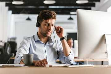 Wall Mural - photo of stressed man 30s wearing office clothes and headset, sitting by computer and working in cal