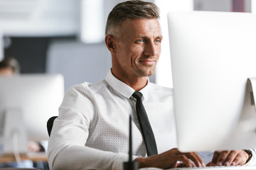 Wall Mural - Image of european businesslike man 30s wearing white shirt and tie sitting at desk in office, and working at computer