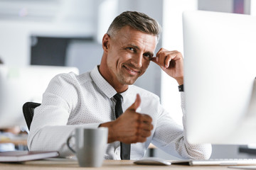 Wall Mural - image of joyful businessman 30s wearing white shirt and tie sitting at desk in office by computer, a