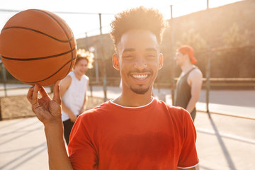 Wall Mural - Image of sporty young boy spinning ball on his finger, while playing basketball at the playground outdoor with his team