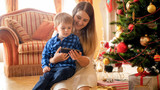 Fototapeta Panele - Portrait of smiling young mother embracing her little boy and watching video on smartphone next to Christmas tree