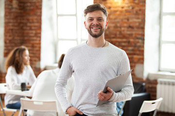 Wall Mural - Confident team leader. Confident young man keeping arms crossed and looking at camera with smile while his colleagues working in the background.