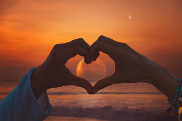 silhouette of couple making heart - shape symbol with moon rising from ocean / sea horizon.