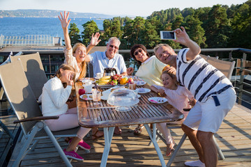 Big happy family having breakfast outdoors on terrace together, sitting around table, drinking coffee. Father taking selfie on phone. Beautiful sea view, warm summer morning. Family portrait.