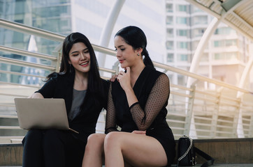 two young business woman in suit talking and reading information about finance news in laptop computer in modern city, financial, investment, teamwork, company success and business growth concept