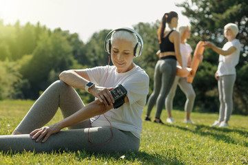 Rhythmical training. Joyful elderly woman sitting on the grass and choosing music before starting her workout in a local park.