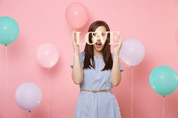 Portrait of surprised happy young woman in blue dress holding wooden word letters love on pastel pink background with colorful air balloons. Birthday holiday party, people sincere emotions concept.