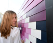 pretty young smiling blonde woman, frame left, looking over shoulder, wearing white shirt, next to receding wall siding purple painted in geometric hues 