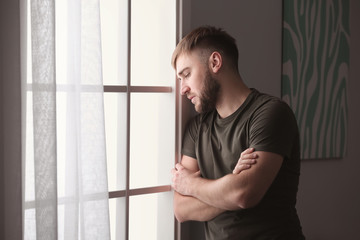 Depressed young man near window at home