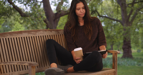 Wall Mural - Millennial woman sitting on park bench using tablet computer and drinking coffee