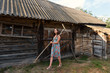 a young woman in a summer retro sarafan dress goes around the yard along the sheds and old rural buildings with rakes on the shoulder