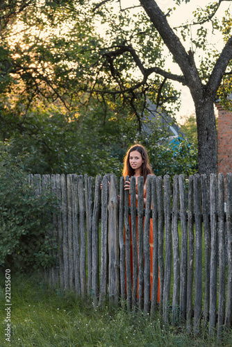 Fotografie, Obraz a sad expectant girl stands behind a wooden palisade in a village attire