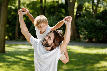 Summer day. Smiling handsome man with dark hairs and beard is holding little boy on his back.