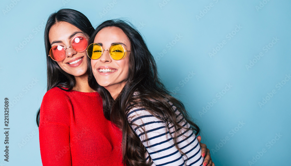 Like a sisters! Two beautiful young girl friends in sunglasses posing with smile and have a fun on blue background - obrazy, fototapety, plakaty 