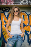 Fototapeta  - Portrait of young beautiful woman wearing white tank shirt and blue jeans on brick wall with graffiti background