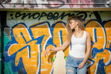 Fototapeta  - Portrait of young beautiful woman wearing white tank shirt and blue jeans on brick wall with graffiti background