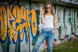 Fototapeta  - Portrait of young woman wearing white shirt and blue jeans and sunglasses on a brick wall with graffiti painting background