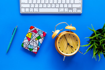 Deadline concept. Planning the day. Notebook and alarm clock on office desk with computer keyboard on blue background top view