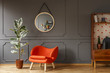 Bright orange armchair, a retro wooden cabinet and a mirror on a gray wall with molding in a stylish living room interior with copy space place for a lamp. Real photo.
