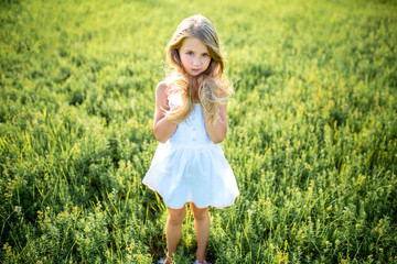 Wall Mural - cute little child in white dress posing in green field and looking at camera