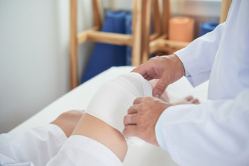 Wall Mural - Anonymous medical practitioner using clean bandage to dress knee of unrecognizable man on blurred background of doctor's office