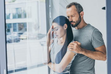 Wall Mural - bearded man supporting sad stressed woman with closed eyes standing near window