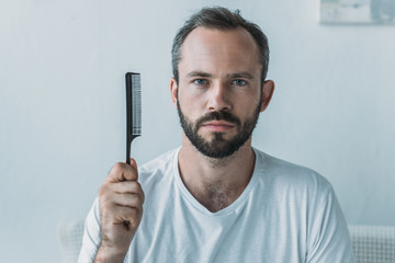 bearded mid adult man holding comb and looking at camera, hair loss concept