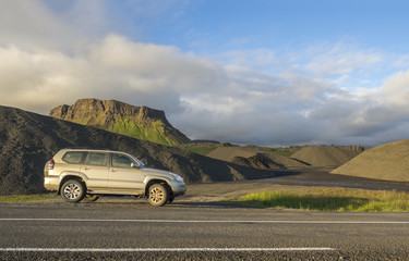 dirty off road car standing next asphalt road with Iceland northern summer landscape, green eroded mountains and blue sky, white clouds