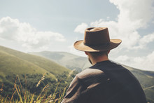 Stylish Traveler Man In Hat Sitting On Top Of Sunny Mountains. Travel And Wanderlust Concept. Space For Text. Happy Hipster Traveling And Camping. Summer Vacation
