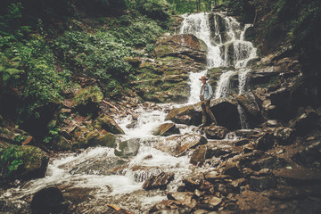 Wall Mural - stylish hipster man in hat with photo camera, standing at waterfall in forest in mountains. traveler guy exploring woods. travel and wanderlust concept. atmospheric moment