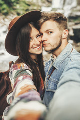 Wall Mural - stylish hipster couple taking selfie and smiling at waterfall in forest in mountains. handsome man and happy woman in hat embracing on honeymoon. travel together. summer vacation