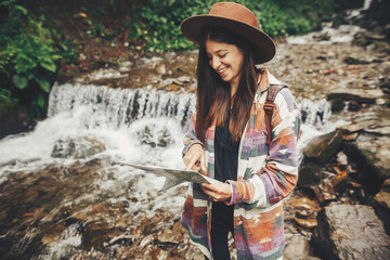 stylish hipster girl in hat with backpack exploring map, standing at river with waterfall in mountains. traveler woman traveling in woods. travel and wanderlust concept. atmospheric moment