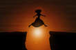 © PixlMakr - Woman wearing a dress and high heels jumps over a gap between two rocks during sunset