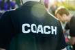 Sport coach in black shirt with white Coach text on the back standing outdoor at a school field, with morning lens flare